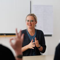 Das Bild zeigt Paulina Pfau beim Gebärden. Sie trägt eine Brille mit sieht zu einer Person, die unscharf im Vordergrund des Bildes sitzt und mit ihrer Hand ein “F” gebärdet.