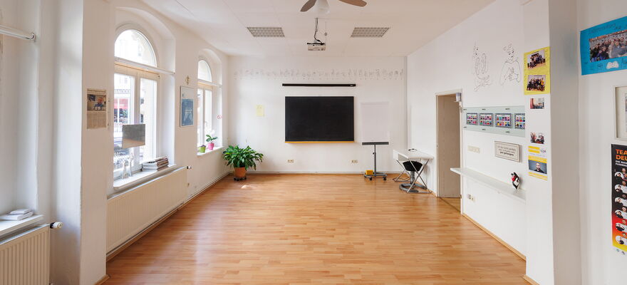 Das Bild zeigt den leeren Kursraum. Er ist weiß mit hellem Parkettboden. Am hinteren Ende befindet sich in der Mitte ein großer Bildschirm. An den Wänden hängen Abbildungen von verschiedenen Gebärden.