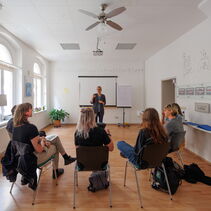 Auf dem Bild ist der Kursraum zu sehen. Fünf Teilnehmer sitzen auf Stühlen und schauen ihre Lehrerin Paulina Pfau an.