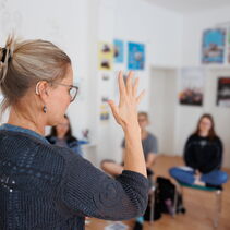 Auf dem Bild ist Paulina Pfau bei einem Gebärdenkurs zu sehen. Ihre Hände sind gespreizt, an ihrem Ohr erkennt man ein Hörgerät. Im Hintergrund sind Kursteilnehmer zu sehen.